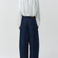 A person is shown from the back, wearing the Simkhai Frida Top—a fluid silhouette long-sleeve white silk georgette blouse featuring blouson sleeves and a buttoned back—paired with wide-leg dark blue jeans. The background is plain and light-colored.