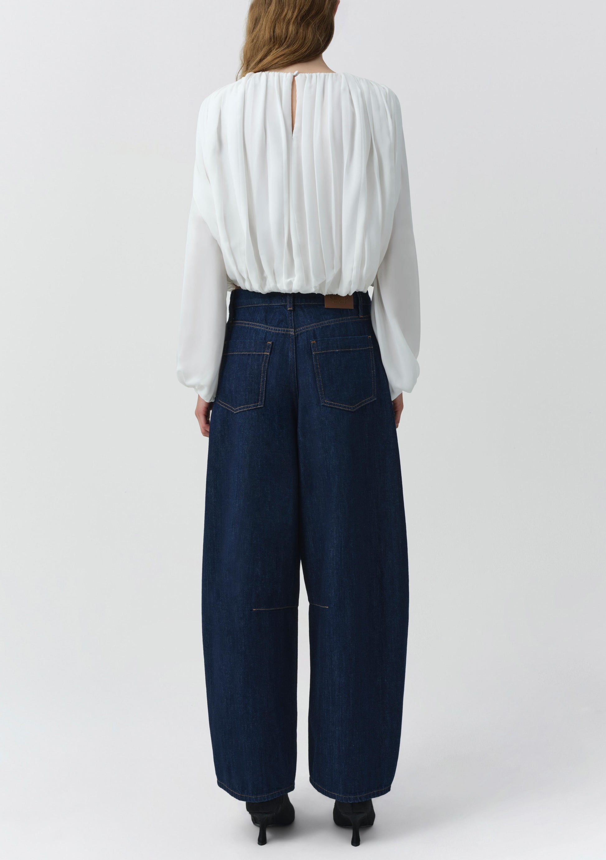 A person is shown from the back, wearing the Simkhai Frida Top—a fluid silhouette long-sleeve white silk georgette blouse featuring blouson sleeves and a buttoned back—paired with wide-leg dark blue jeans. The background is plain and light-colored.