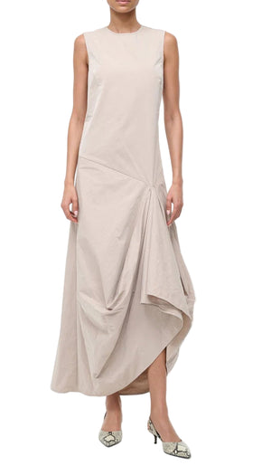 A woman wears the Staud Viola Dress, a sleeveless light beige piece with an asymmetrical hem and draped skirt, styled with pointed-toe snakeskin heels.