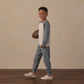 A young boy stands on a wooden floor, smiling at the camera with a football in hand against a plain beige background, enjoying breathable comfort in every moment in the Rylee & Cru Boys’ Long Sleeve Raglan Tee.