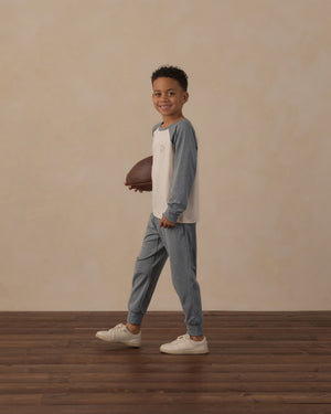 A young boy stands on a wooden floor, smiling at the camera with a football in hand against a plain beige background, enjoying breathable comfort in every moment in the Rylee & Cru Boys’ Long Sleeve Raglan Tee.