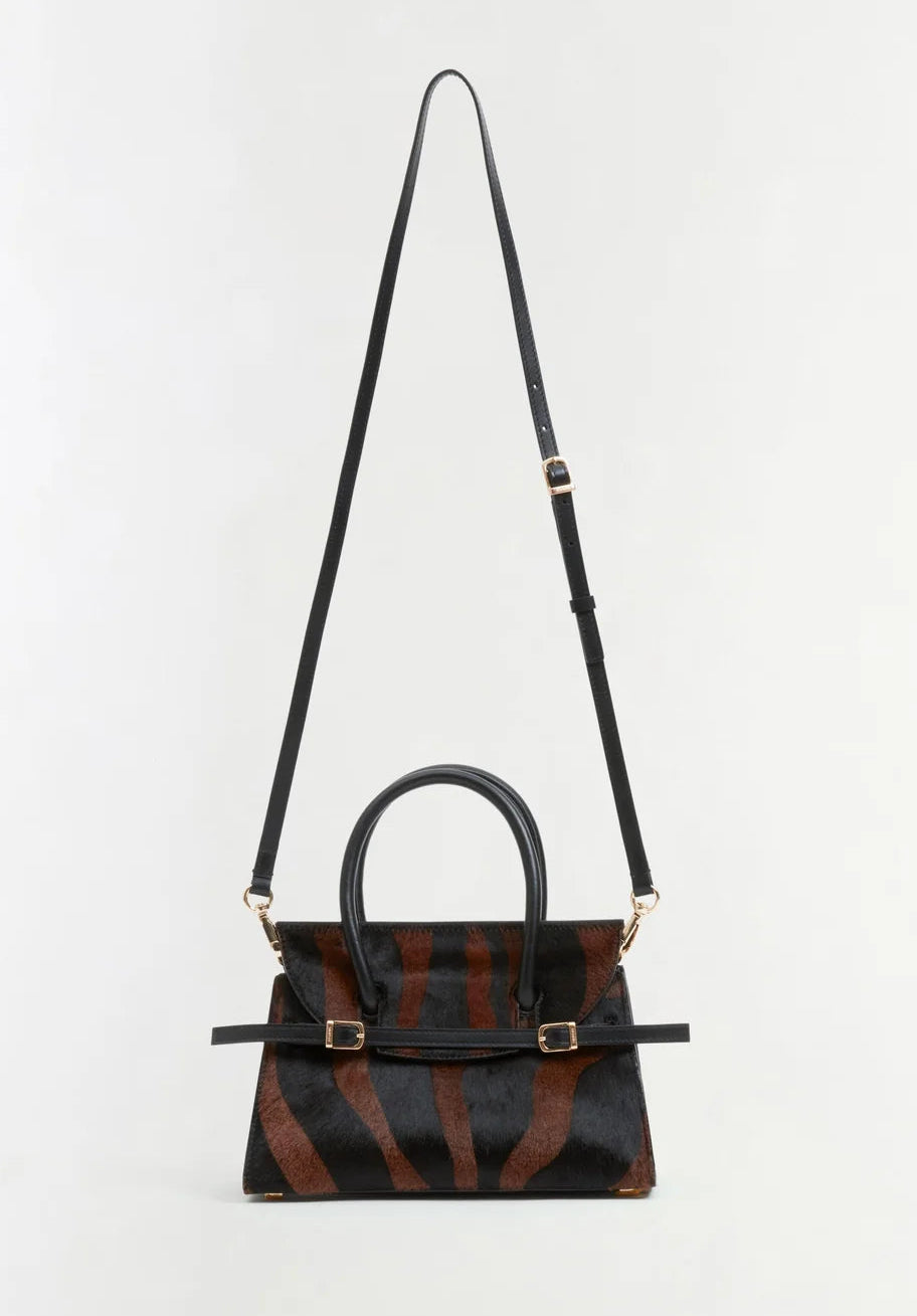 The Simkhai River Small Top Handle Bag is styled in a bold black and brown tiger-print pattern, enhanced by gold hardware and a long adjustable crossbody strap, and is displayed against a plain light background.