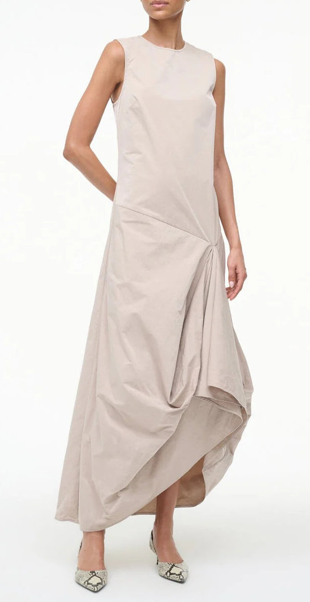 A woman wearing the Staud Viola Dress—sleeveless with a draped skirt and asymmetrical hem—pairs it with pointed-toe snakeskin flats while standing against a plain white background.