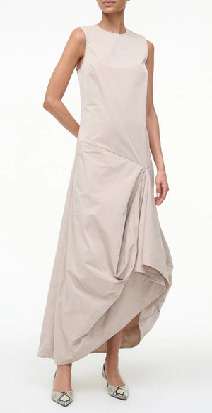 A woman wearing the Staud Viola Dress—sleeveless with a draped skirt and asymmetrical hem—pairs it with pointed-toe snakeskin flats while standing against a plain white background.