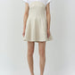A woman faces forward wearing the Simkhai Sundae Linen Mini Dress—a light beige mini dress with an A-line skirt and structured bodice—layered over a white collared short-sleeve shirt and paired with white sandals.