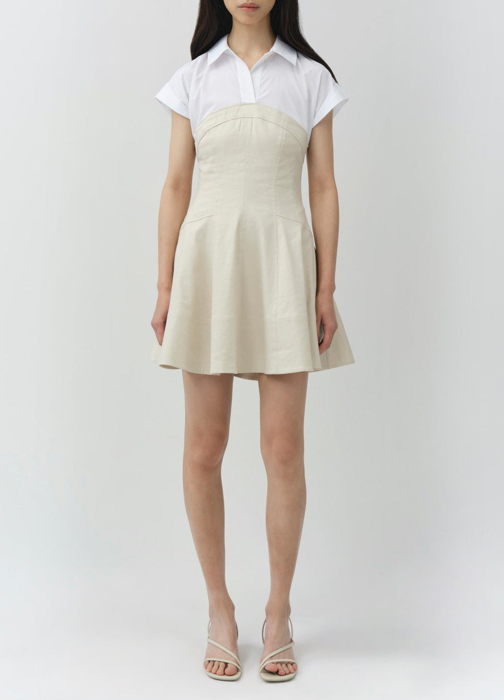 A woman faces forward wearing the Simkhai Sundae Linen Mini Dress—a light beige mini dress with an A-line skirt and structured bodice—layered over a white collared short-sleeve shirt and paired with white sandals.
