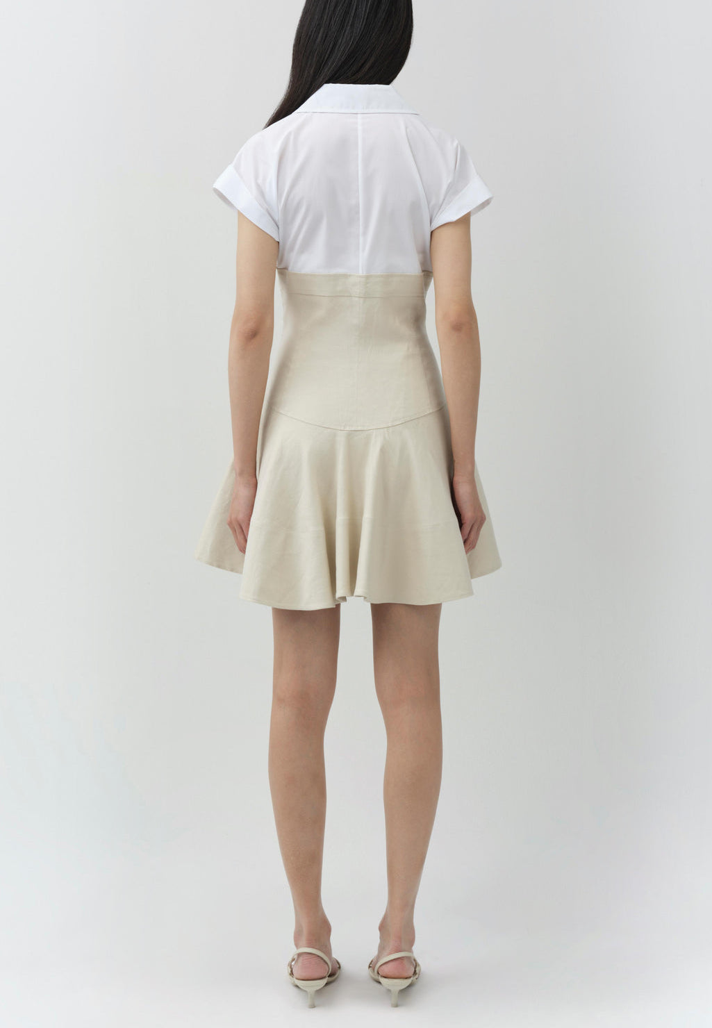A woman faces away in the Simkhai Sundae Linen Mini Dress, featuring a structured bodice and beige flared skirt. Her long dark hair and light-colored heels complete an effortlessly elegant look.