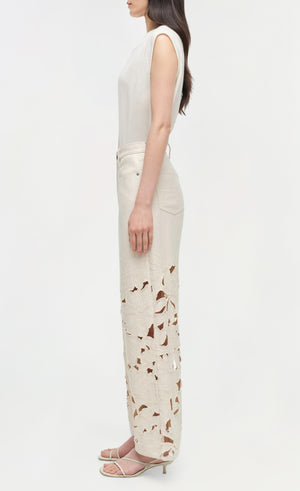 Woman in profile wears Simkhai’s Betty Floral Cut Out Jean—high-waisted straight leg denim with floral embroidery and cutout details—paired with a sleeveless white top and strappy heeled sandals.