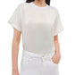A woman wears the Simkhai Addy Satin T-Shirt—a plain white, relaxed-fit short-sleeve top with a round neckline—paired with white jeans and stands with one hand in her pocket.