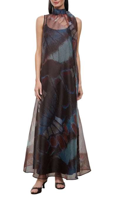 A woman is wearing the Staud Albee Dress, an organza sleeveless design with a high neckline and abstract butterfly wing pattern in brown, blue, and red hues. This elegant dress features a delicate interior slip for comfort and is paired perfectly with black open-toe heels.