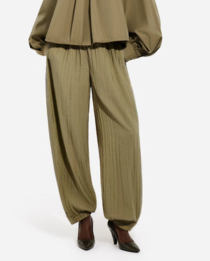 A person wears the CO Articulated Vented Pant in olive green lightweight technical fabric with pleats, elasticated waist and cuffs, paired with black pointed-toe heels. Only the lower half of the outfit is visible.