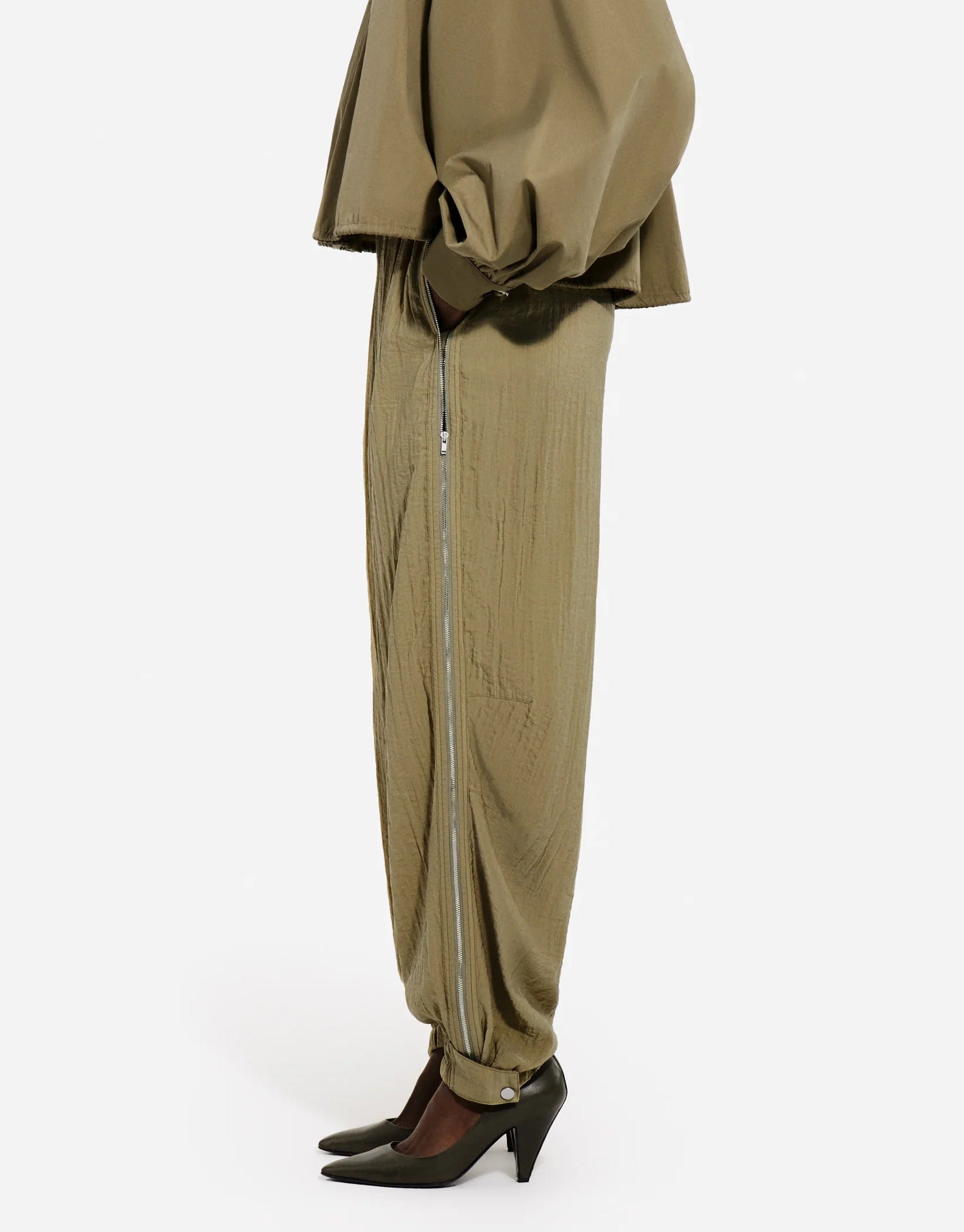A person in side profile wears CO’s olive Articulated Vented Pant with an elastic waist, a matching top, and dark green pointed heels, set against a plain white background.