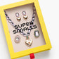 The Lavender Haze Mega Jewelry Set by Super Smalls comes in a yellow box and includes a purple necklace with a pendant, plus two pairs of earrings featuring heart and gem designs.