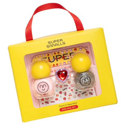 A yellow "Super Smalls" box displays the Paint it Heartfelt Mini Nail Kit with two glitter polishes, a glitter pot, heart nail stickers, and a heart-shaped ring seen through its clear window.