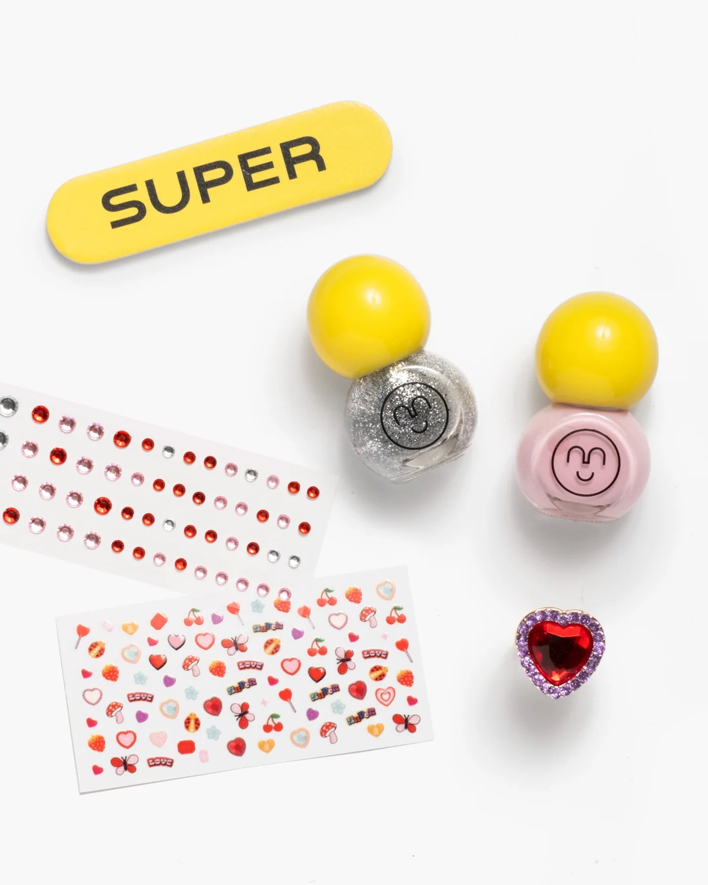 The Paint it Heartfelt Mini Nail Kit by Super Smalls features a yellow “SUPER” nail file, two glitter polishes, decorative and heart stickers, gem stickers, and a heart-shaped ring—all arranged on a white background.