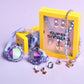 The Lavender Haze Mega Jewelry Set by Super Smalls, with rings, earrings, a purple necklace, and two yellow branded boxes, is displayed on a light purple surface alongside colorful swim goggles.