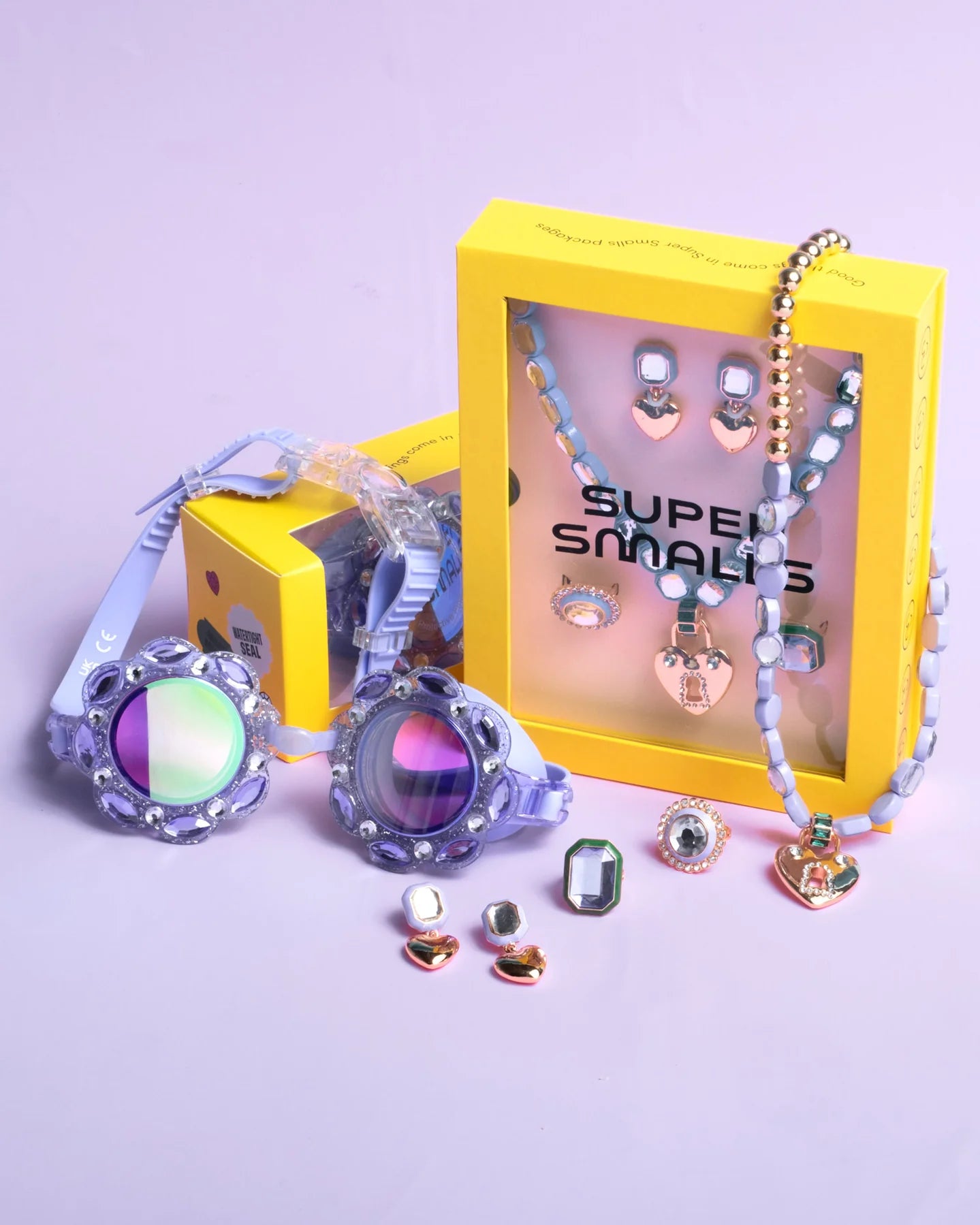 The Lavender Haze Mega Jewelry Set by Super Smalls, with rings, earrings, a purple necklace, and two yellow branded boxes, is displayed on a light purple surface alongside colorful swim goggles.