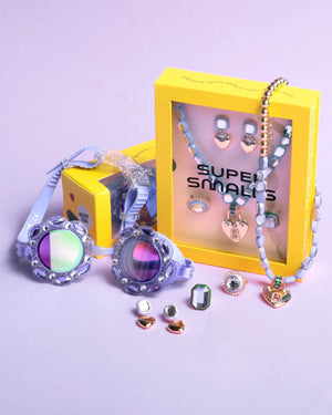 The Lavender Haze Mega Jewelry Set by Super Smalls, with rings, earrings, a purple necklace, and two yellow branded boxes, is displayed on a light purple surface alongside colorful swim goggles.