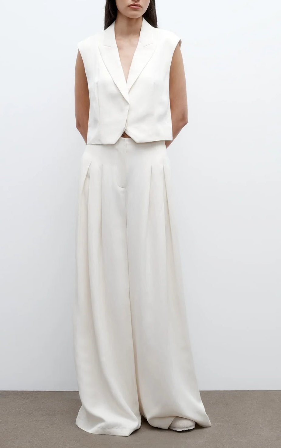 A woman poses against a plain background in TWP’s Drew Coated Viscose Linen Pant, styled with an off-white sleeveless blazer; the matching high-rise, wide-leg pants offer a sleek look with her arms behind her back.