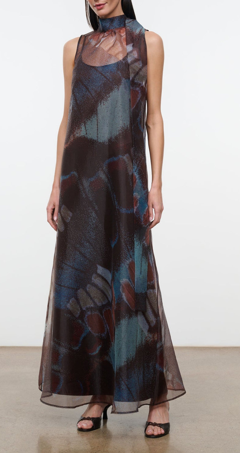 A woman wearing the Staud Albee Dress, an organza sleeveless gown with a floor-length, sheer silhouette and an abstract blue, brown, and red pattern, styled with black sandals and finished with an interior slip for coverage.