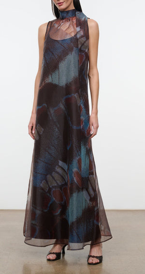 A woman wearing the Staud Albee Dress, an organza sleeveless gown with a floor-length, sheer silhouette and an abstract blue, brown, and red pattern, styled with black sandals and finished with an interior slip for coverage.