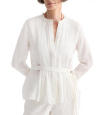 A woman wears the TWP Pleated Poet Blouse, a white linen top with gathered detailing, paired with matching pants. She stands with hands on hips against a plain white background.