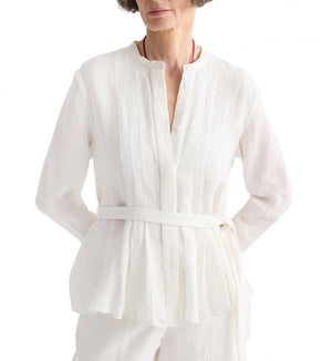 A woman wears the TWP Pleated Poet Blouse, a white linen top with gathered detailing, paired with matching pants. She stands with hands on hips against a plain white background.