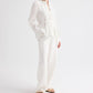 A person wearing the TWP Pleated Poet Blouse—a white linen top with gathered detailing and a belt—paired with matching white wide-leg pants, standing against a plain white background and wearing beige sandals.