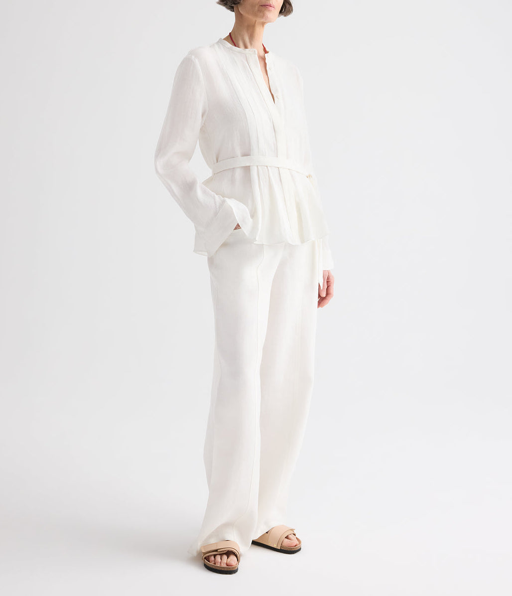 A person wearing the TWP Pleated Poet Blouse—a white linen top with gathered detailing and a belt—paired with matching white wide-leg pants, standing against a plain white background and wearing beige sandals.