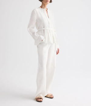 A person wearing the TWP Pleated Poet Blouse—a white linen top with gathered detailing and a belt—paired with matching white wide-leg pants, standing against a plain white background and wearing beige sandals.