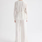 A person stands facing away against a plain white background, wearing the TWP Pleated Poet Blouse— a long-sleeved, semi-sheer belted blouse with gathered detailing— paired with wide-leg white trousers.