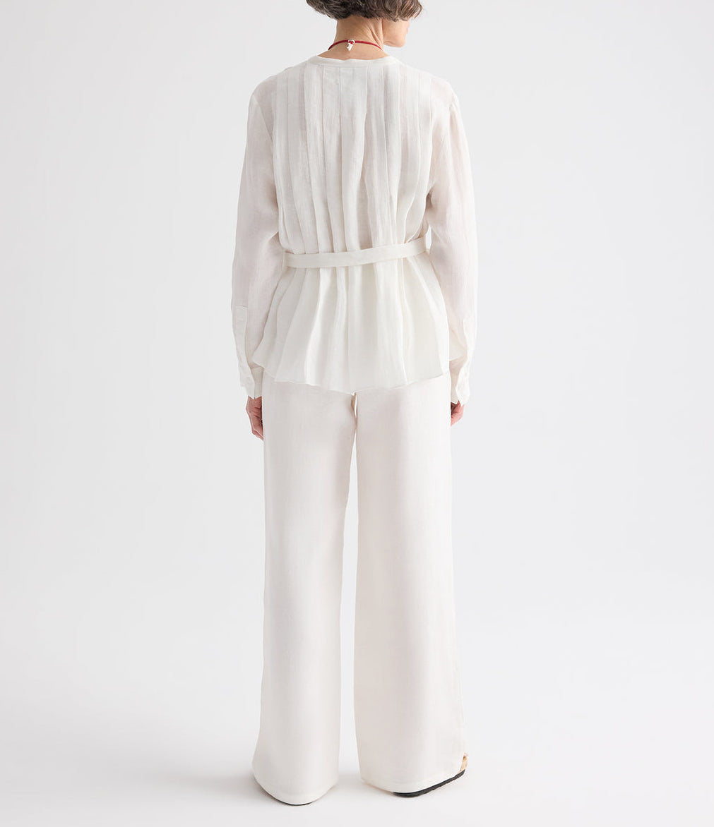 A person stands facing away against a plain white background, wearing the TWP Pleated Poet Blouse— a long-sleeved, semi-sheer belted blouse with gathered detailing— paired with wide-leg white trousers.