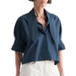 A woman wearing the TWP Tie One On Shirt in navy blue Supima cotton with short sleeves and a wide collar, paired with white pants, stands against a white background.