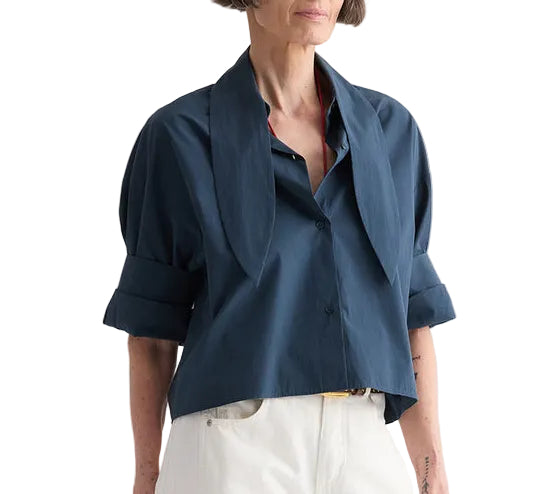 A woman wearing the TWP Tie One On Shirt in navy blue Supima cotton with short sleeves and a wide collar, paired with white pants, stands against a white background.