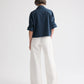 A person stands facing away from the camera against a plain white background, wearing TWP's Tie One On Shirt—a vintage-washed poplin dark blue long-sleeve shirt with rolled-up sleeves—paired with loose-fitting white pants.