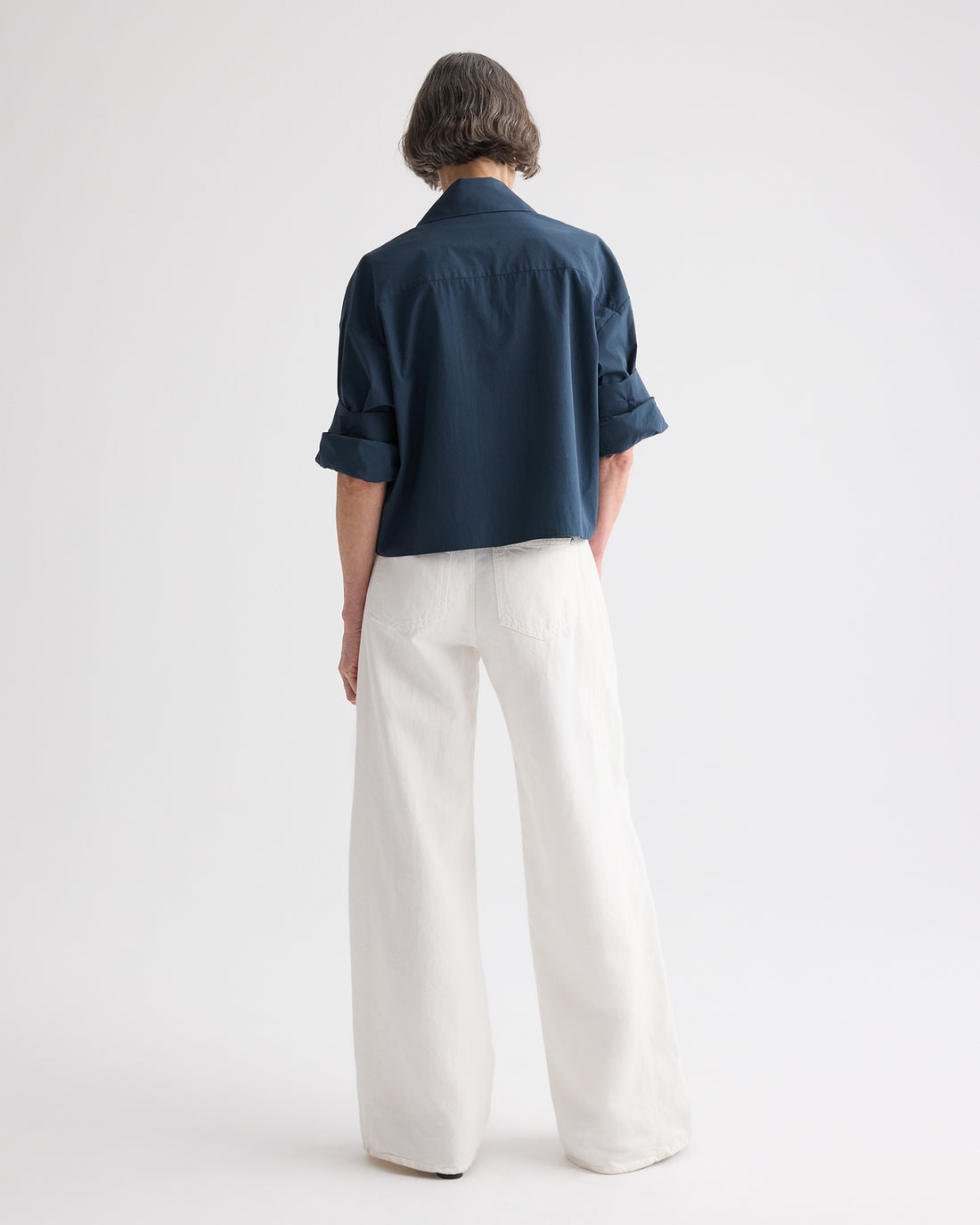 A person stands facing away from the camera against a plain white background, wearing TWP's Tie One On Shirt—a vintage-washed poplin dark blue long-sleeve shirt with rolled-up sleeves—paired with loose-fitting white pants.