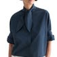 A person wearing the TWP Tie One On Shirt—a dark blue, vintage-washed poplin button-up blouse featuring a large necktie detail and rolled-up sleeves—stands against a white background.