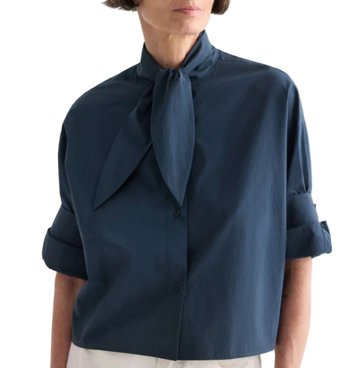 A person wearing the TWP Tie One On Shirt—a dark blue, vintage-washed poplin button-up blouse featuring a large necktie detail and rolled-up sleeves—stands against a white background.