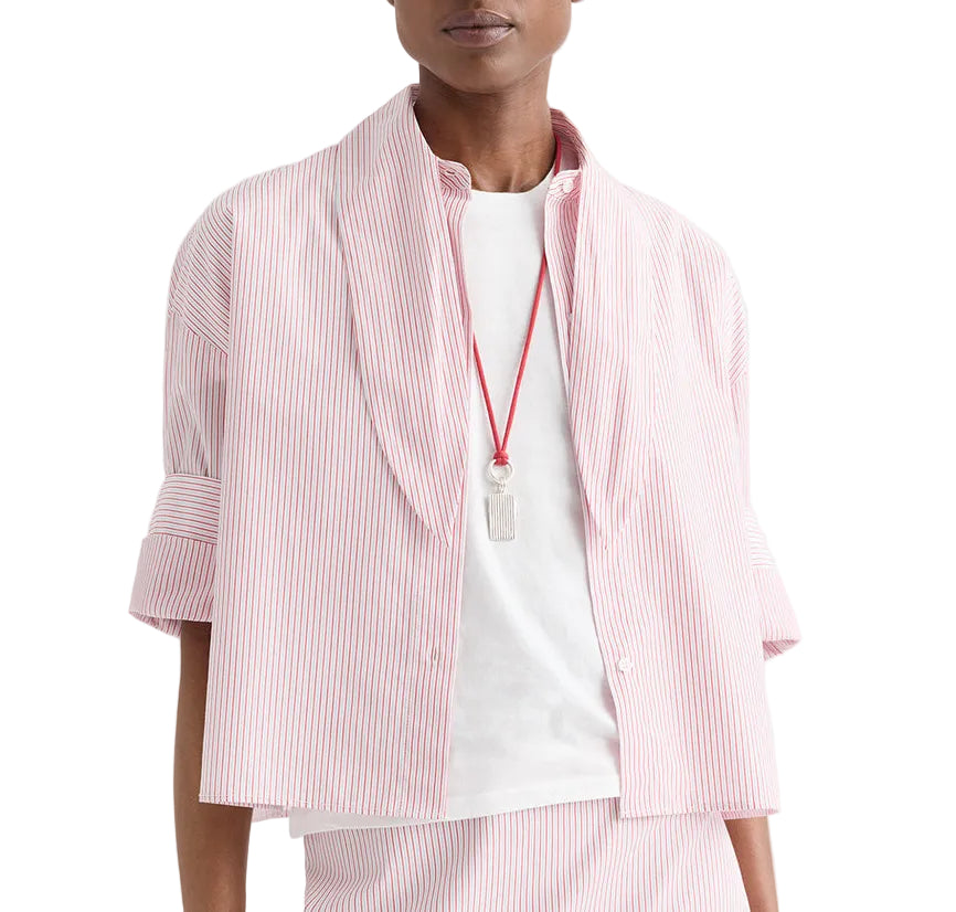 A person wearing the TWP Tie One On Stripe Shirt—a pink and white vertically striped button-up with rolled-up sleeves—layered over a white T-shirt, accessorized with a clear pendant on a red cord necklace.