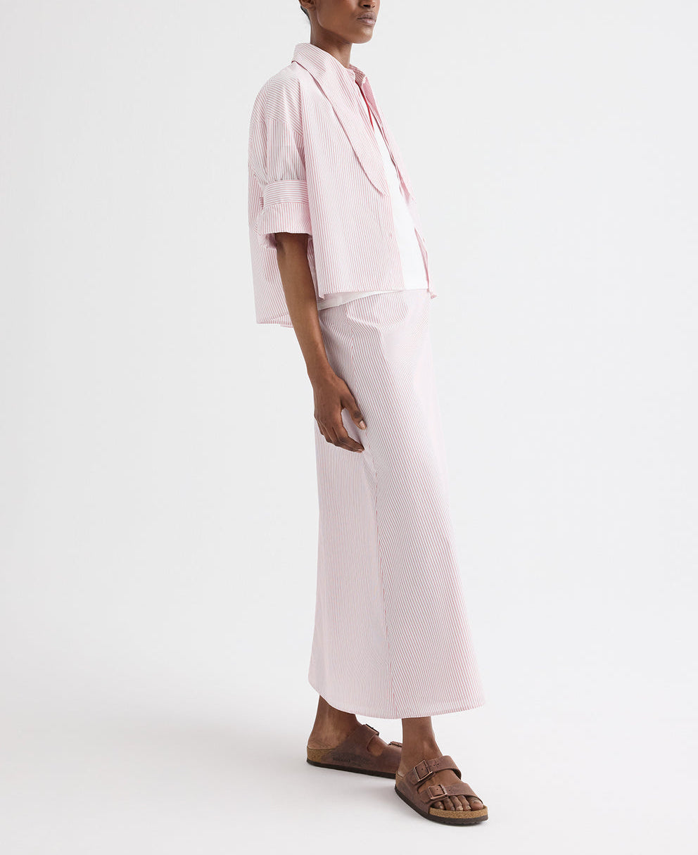 A person wearing the TWP Tie One On Stripe Shirt—a light pink, collared, cotton short-sleeve button-up with stripes—paired with a matching long skirt and brown sandals, stands against a plain white background.