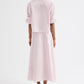 A person stands facing away against a plain white background, wearing the TWP Tie One On Stripe Shirt in light pink with rolled cuffs and subtle cotton stripes, paired with a matching ankle-length skirt and brown sandals.