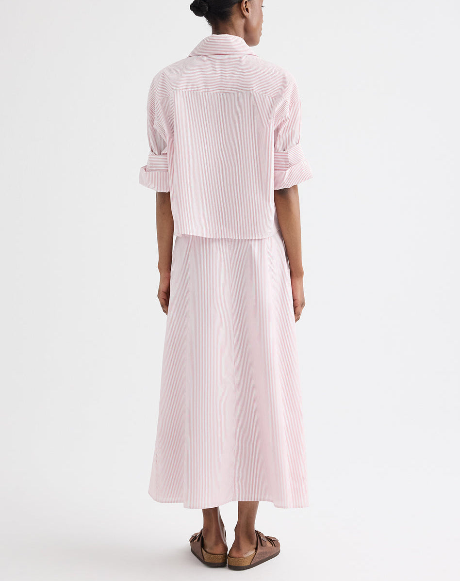A person stands facing away against a plain white background, wearing the TWP Tie One On Stripe Shirt in light pink with rolled cuffs and subtle cotton stripes, paired with a matching ankle-length skirt and brown sandals.