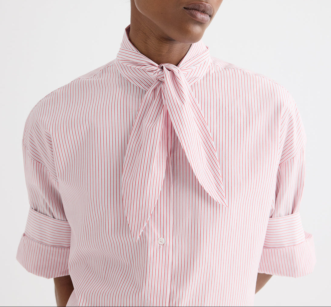Model wears the TWP Tie One On Stripe Shirt, a cropped short-sleeve button-down featuring red cotton stripes and a necktie knot detail, with rolled sleeves, shown from shoulders to waist against a plain background.