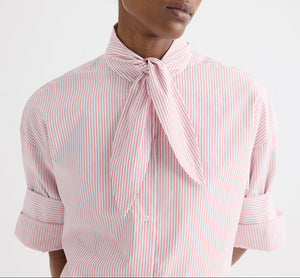 Model wears the TWP Tie One On Stripe Shirt, a cropped short-sleeve button-down featuring red cotton stripes and a necktie knot detail, with rolled sleeves, shown from shoulders to waist against a plain background.