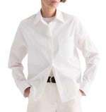 A person wears the TWP Cowboy Update Shirt—a classic white poplin shirt with a front pocket—paired with white pants and a black belt with a gold buckle, hands casually in their pockets.