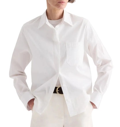 A person wears the TWP Cowboy Update Shirt—a classic white poplin shirt with a front pocket—paired with white pants and a black belt with a gold buckle, hands casually in their pockets.