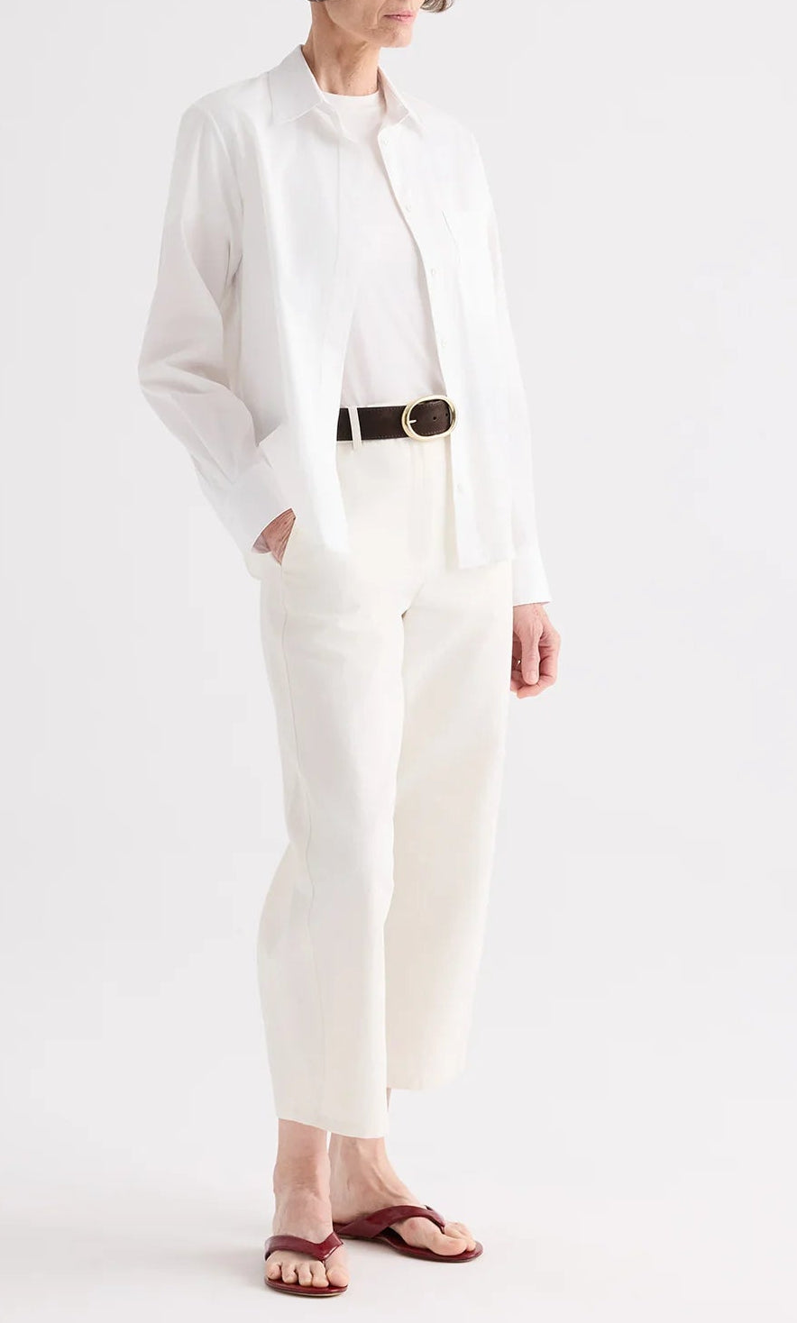 Wearing the TWP Cowboy Update Shirt, a person stands hands in pockets against a plain background, paired with a white t-shirt, white pants, black belt, and brown sandals.