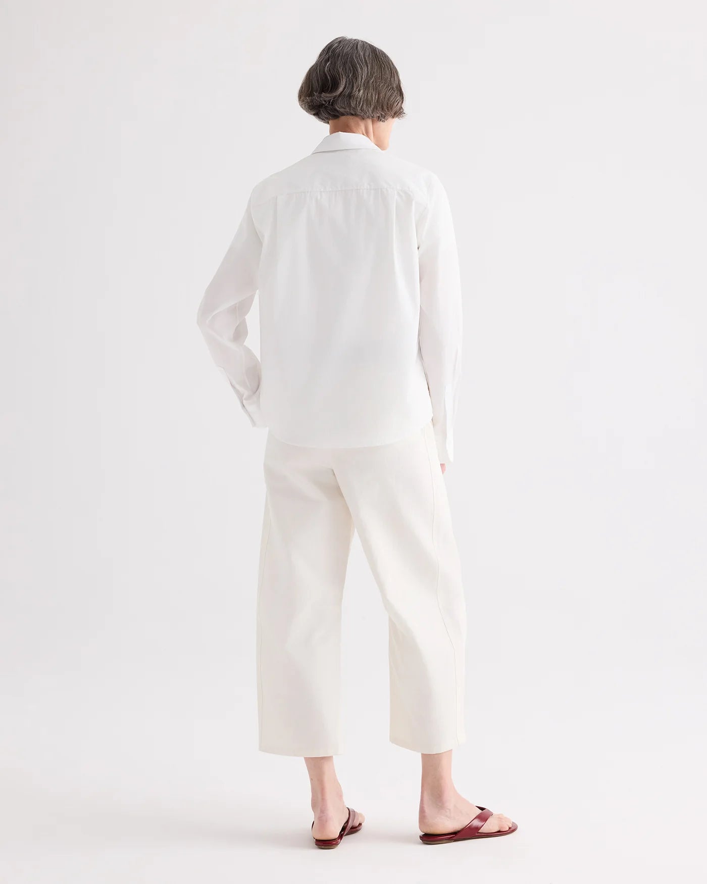 A person with short gray hair stands facing away, wearing the TWP Cowboy Update Shirt and white wide-leg pants with red sandals against a plain white background.