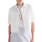 A woman wears the TWP Short Lived Cherry Pleat Top over a white top, paired with a red necklace and a light blue striped skirt, set against a white background.