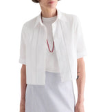 A woman wears the TWP Short Lived Cherry Pleat Top over a white top, paired with a red necklace and a light blue striped skirt, set against a white background.